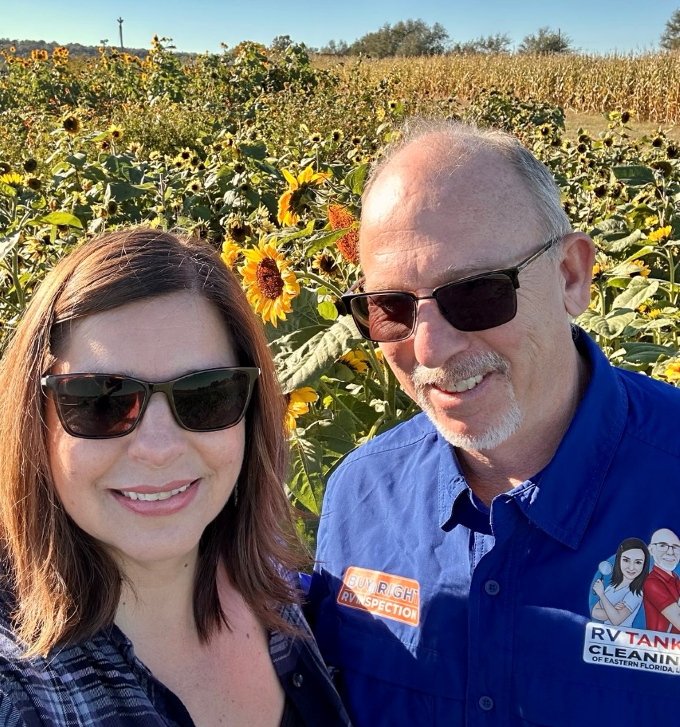 Smiling couple wearing sunglasses posing for a selfie in a sunflowers field on a sunny day.