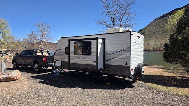 Pickup truck towing a white travel trailer with a slide-out extended, parked on a gravel campsite by a lake and hills in the background