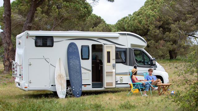 RV parked in a grassy campsite with two surfboards propped against the side and two people relaxing at a small table outside the door.