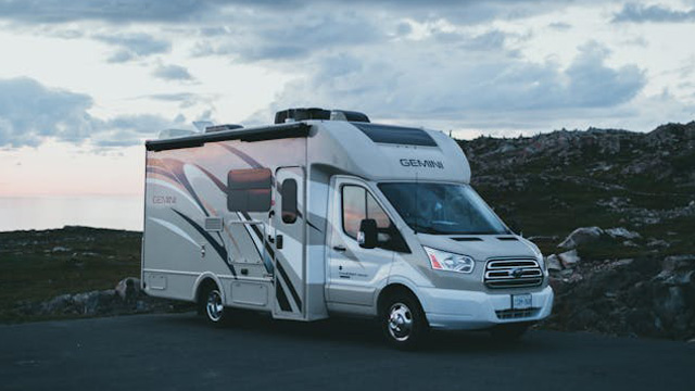 White motorhome parked on a coastal road with rocky hills at dusk, calm sea in the distance under a cloudy sky.