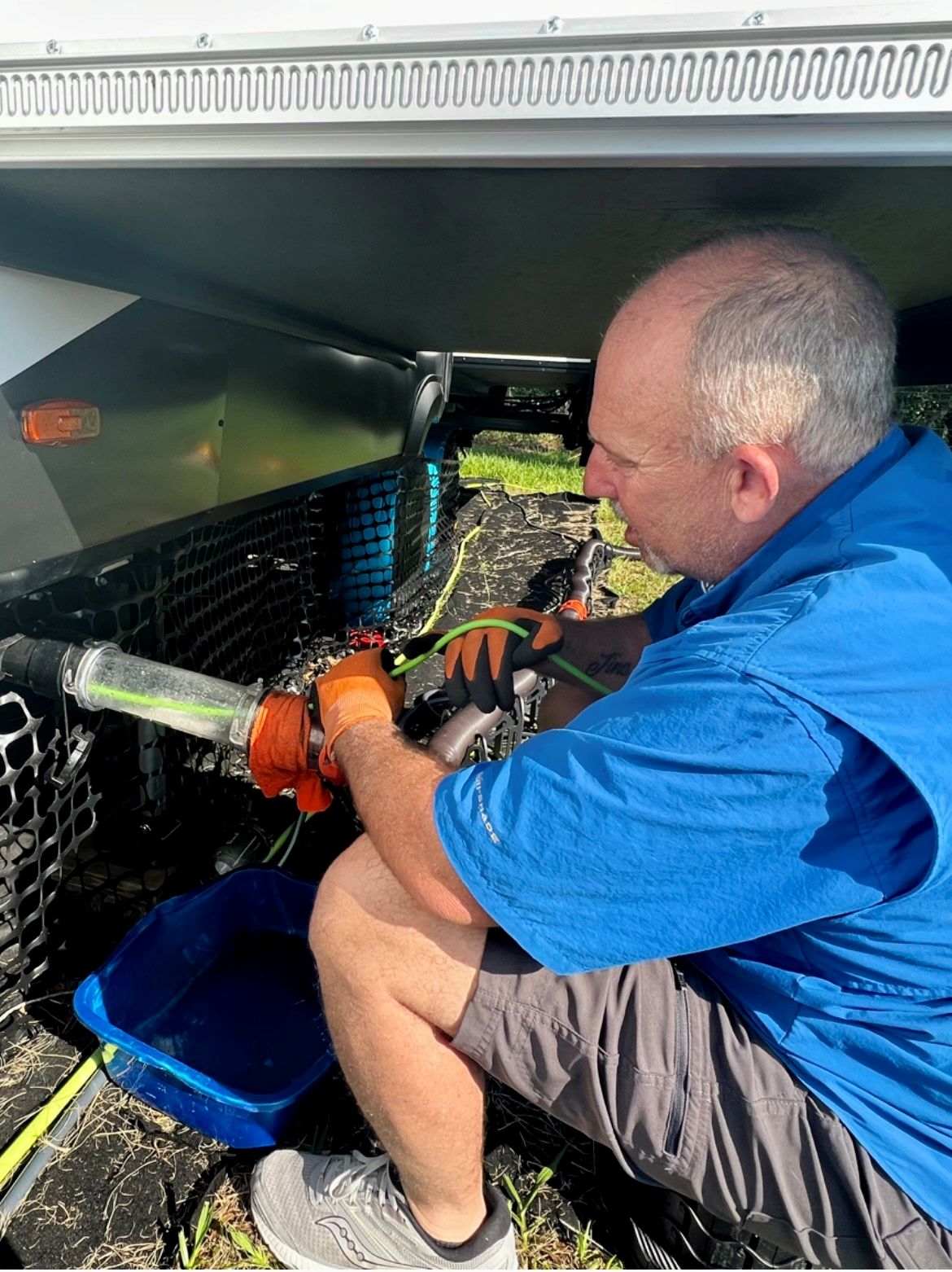 Male technician in a blue shirt kneels beside a vehicle's undercarriage, using a tool to connect or secure hoses near a mesh guard and storage tray.