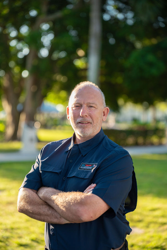 A middle-aged man with a gray beard stands in a sunlit park with arms crossed, wearing a navy uniform with a cleaning company logo.