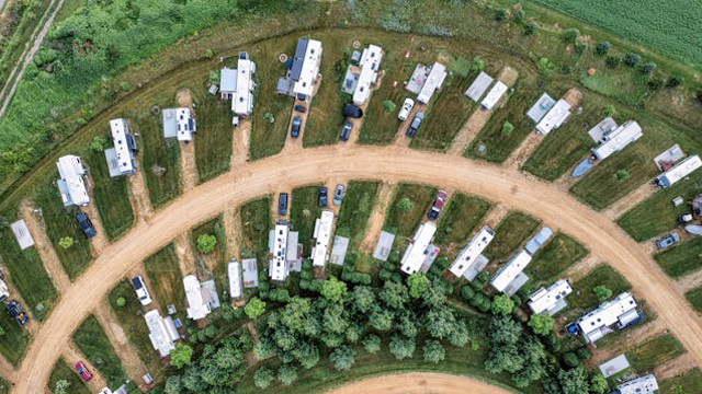 Aerial view of a curved dirt road in an RV park with rows of white mobile homes and parked cars, surrounded by green fields and trees.