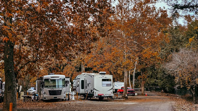 RVs parked at a fall campsite under orange maple trees on a dirt road sunlit by autumn leaves in the park as campers gather nearby.