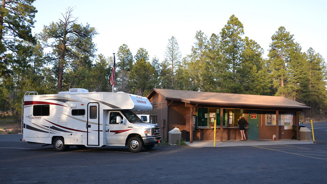White motorhome parked in a forested campground beside a wood-framed check-in building; a person stands at the window.