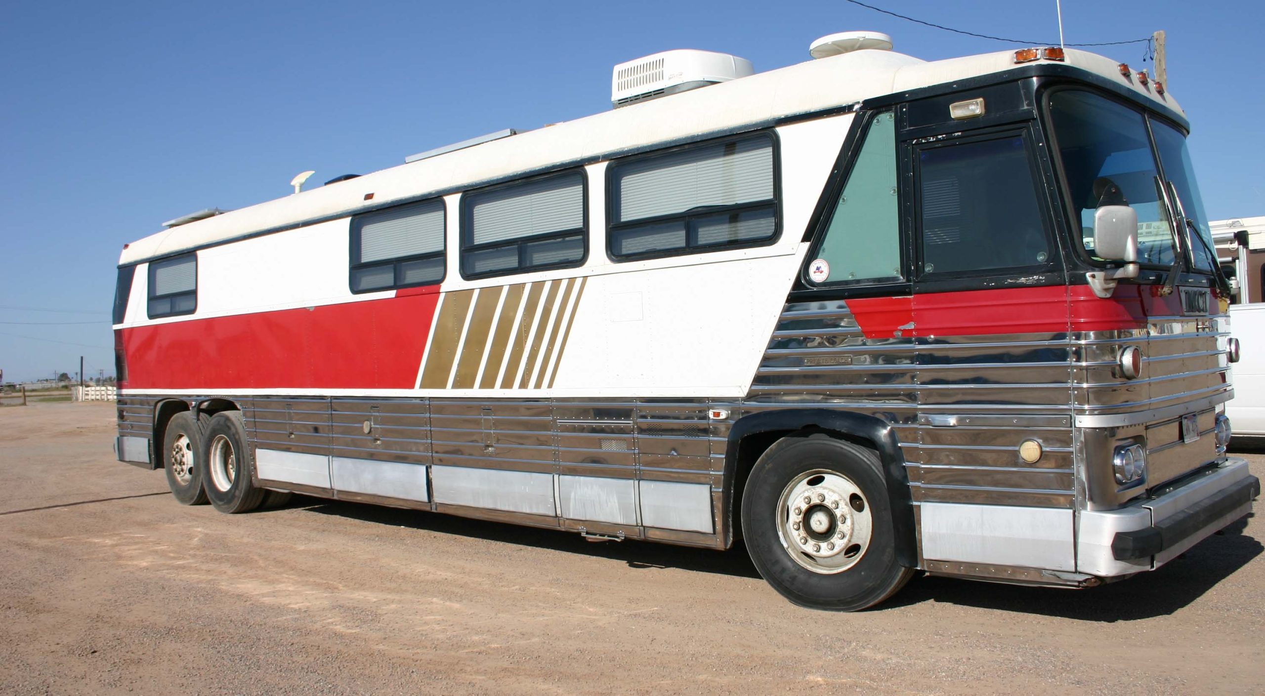 Long retro tour bus with red, white, and chrome exterior parked on a dirt lot under a clear blue sky.
