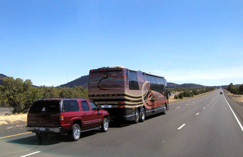 Red SUV towing a large beige-and-brown motorhome on a straight highway with desert hills in the distance and a clear blue sky.