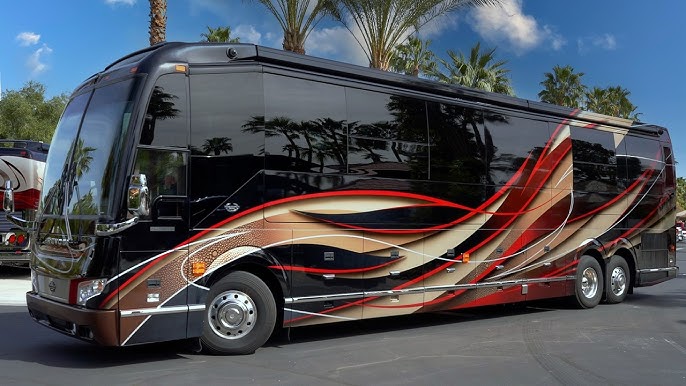 Black luxury tour bus with red, brown, and cream sweeping graphics, parked in a lot with palm trees in the background.