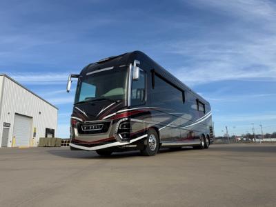 Front-left view of a large black and gray Class A motorhome with red and silver graphics, parked on a sunny concrete lot near a warehouse.