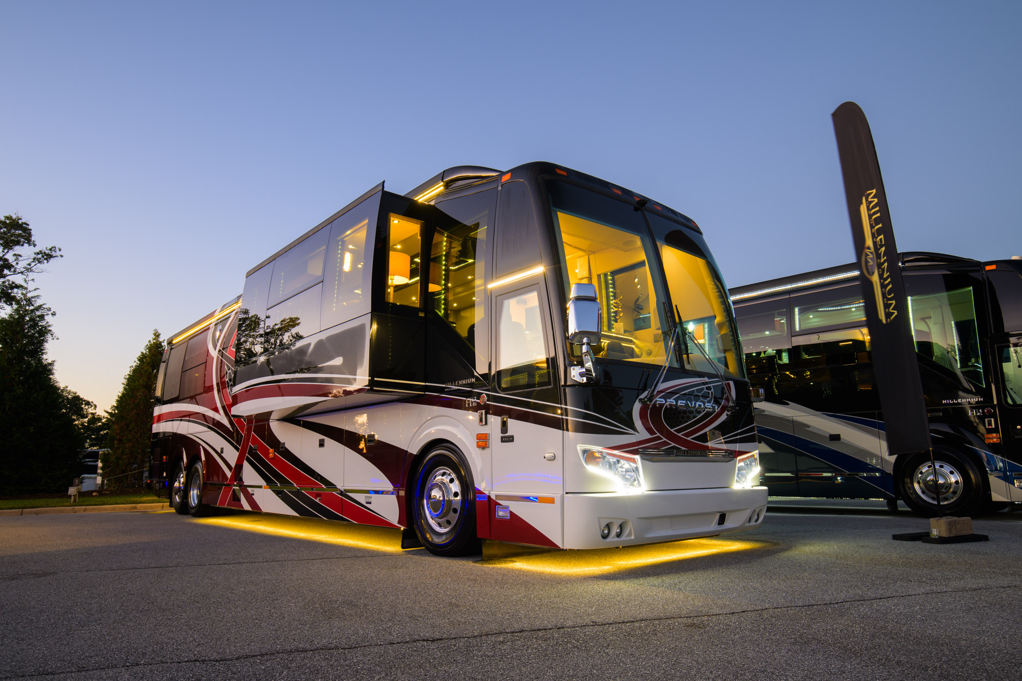 Two luxury tour buses parked side by side at dusk, glossy white with red and black graphics and warm interior lighting under yellow underglow.